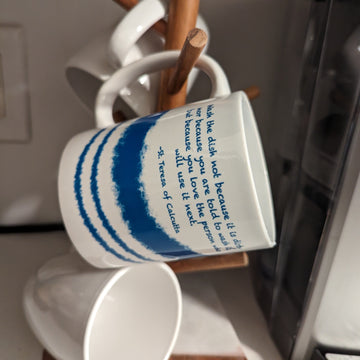 A Catholic mug on a wooden mug hanger that displays Mother Teresa's Wash the Dish quote with blue and white stripes