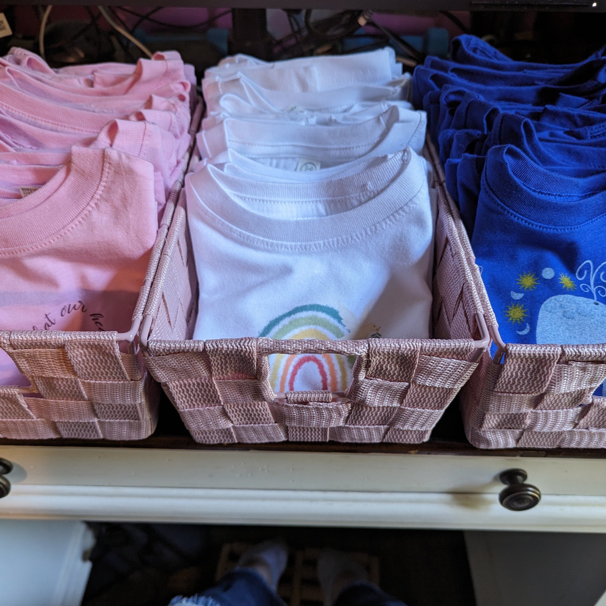 Three baskets of Catholic toddler t-shirts in pink, white, and blue with Catholic symbols on them