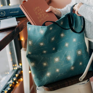 A Catholic tote bag sitting on a chair as a woman puts a devotional into it. The bag is decorated with monstrance illustrations and words from Benediction.